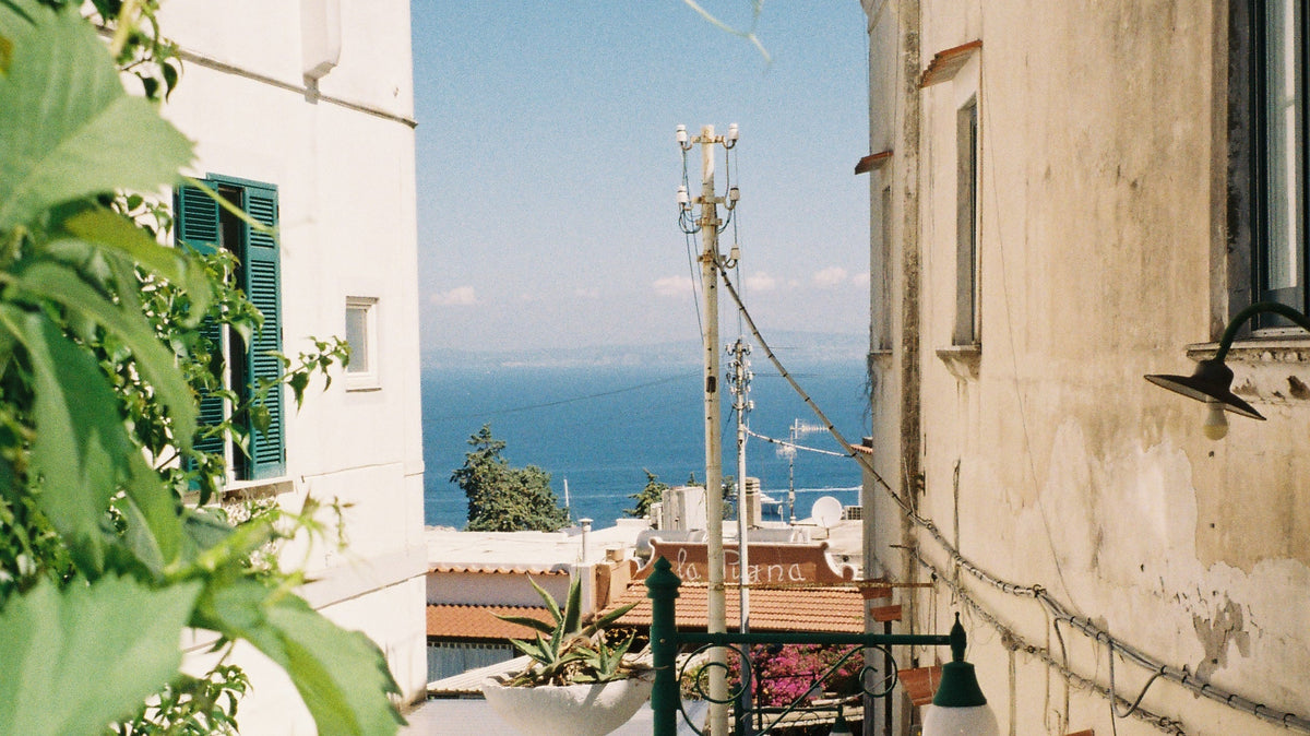 A narrow white alley on Via Mulo, Capri, framed by leaves, with a glimpse of the blue sea at the end