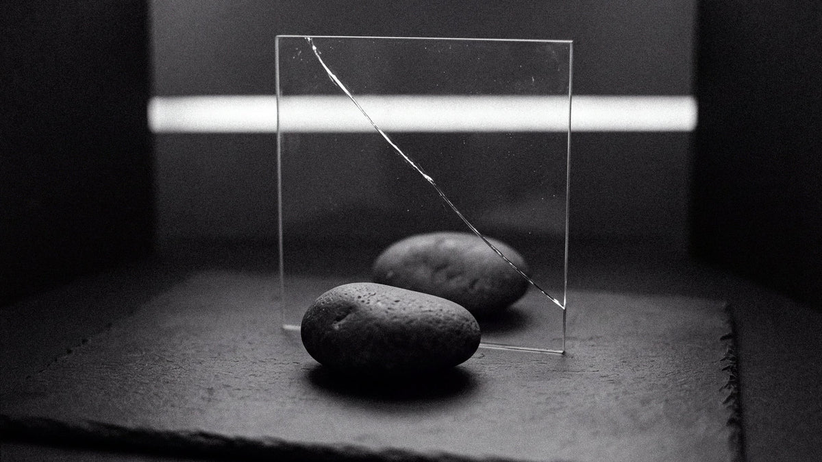 Two smooth dark stones on a slate surface, one on each side of an upright pane of glass with a single crack running through it, a strip of soft light glowing behind them in a dark room.
