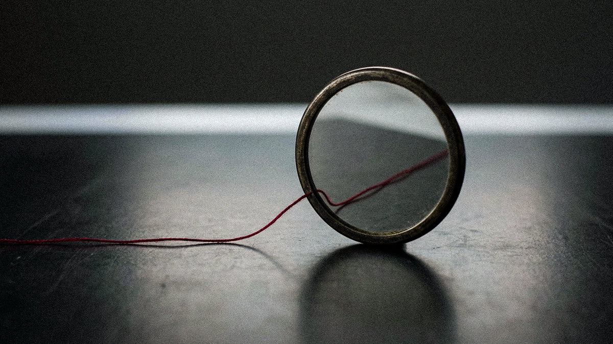 An antique round mirror stands upright on a dark surface, a fine red thread entering its frame and continuing inside the reflection, against a soft band of light.