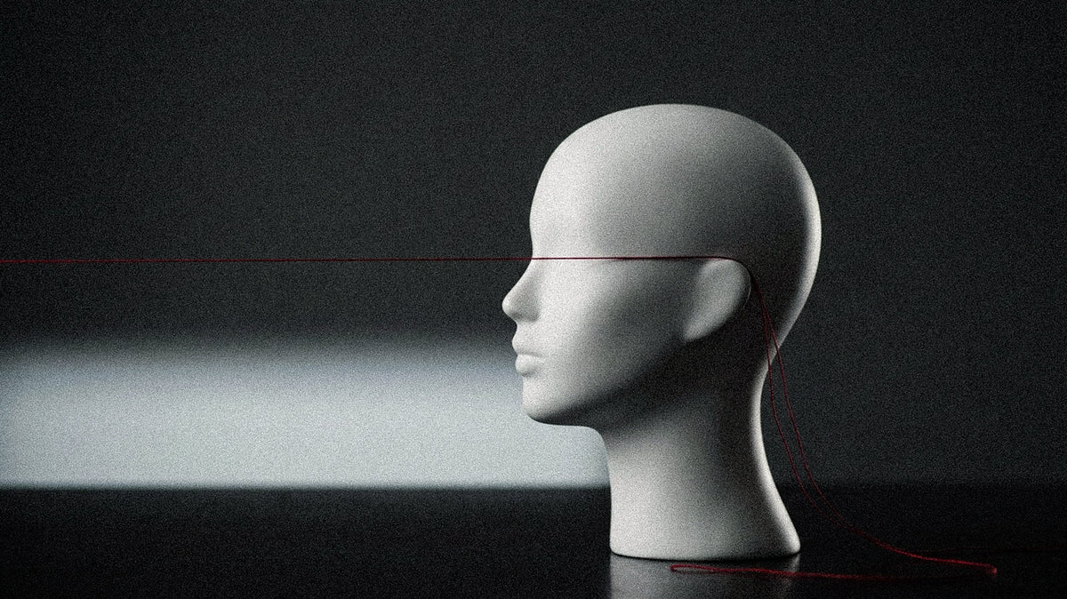 A smooth white mannequin head in profile on a dark surface, a fine red thread running straight across where the eyes would be, its loose ends falling behind the ear.