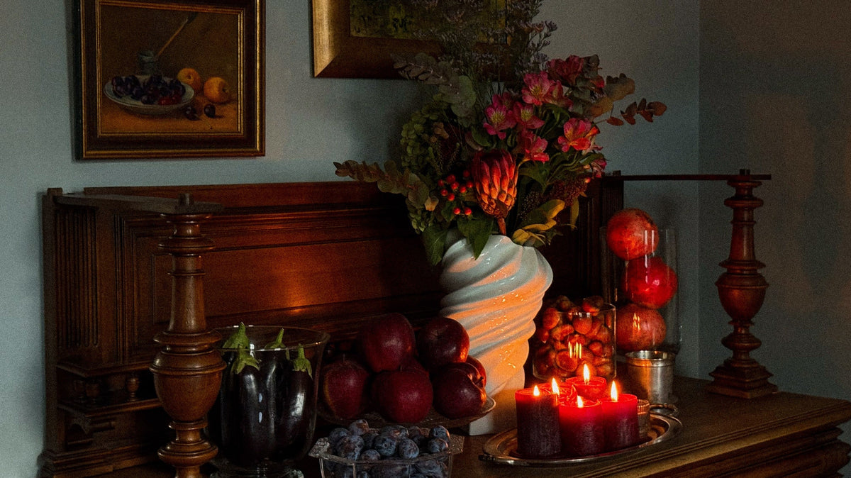 A fall still life on a dark wooden sideboard: a bouquet of alstroemerias and protea in a white twisted vase, red apples, plums in crystal, aubergines, pomegranates, and lit red candles on a silver tray.