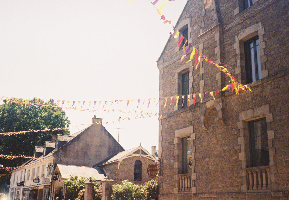 A stone house in Guérande with pink, yellow and violet bunting strung across the street under a pale summer sky.
