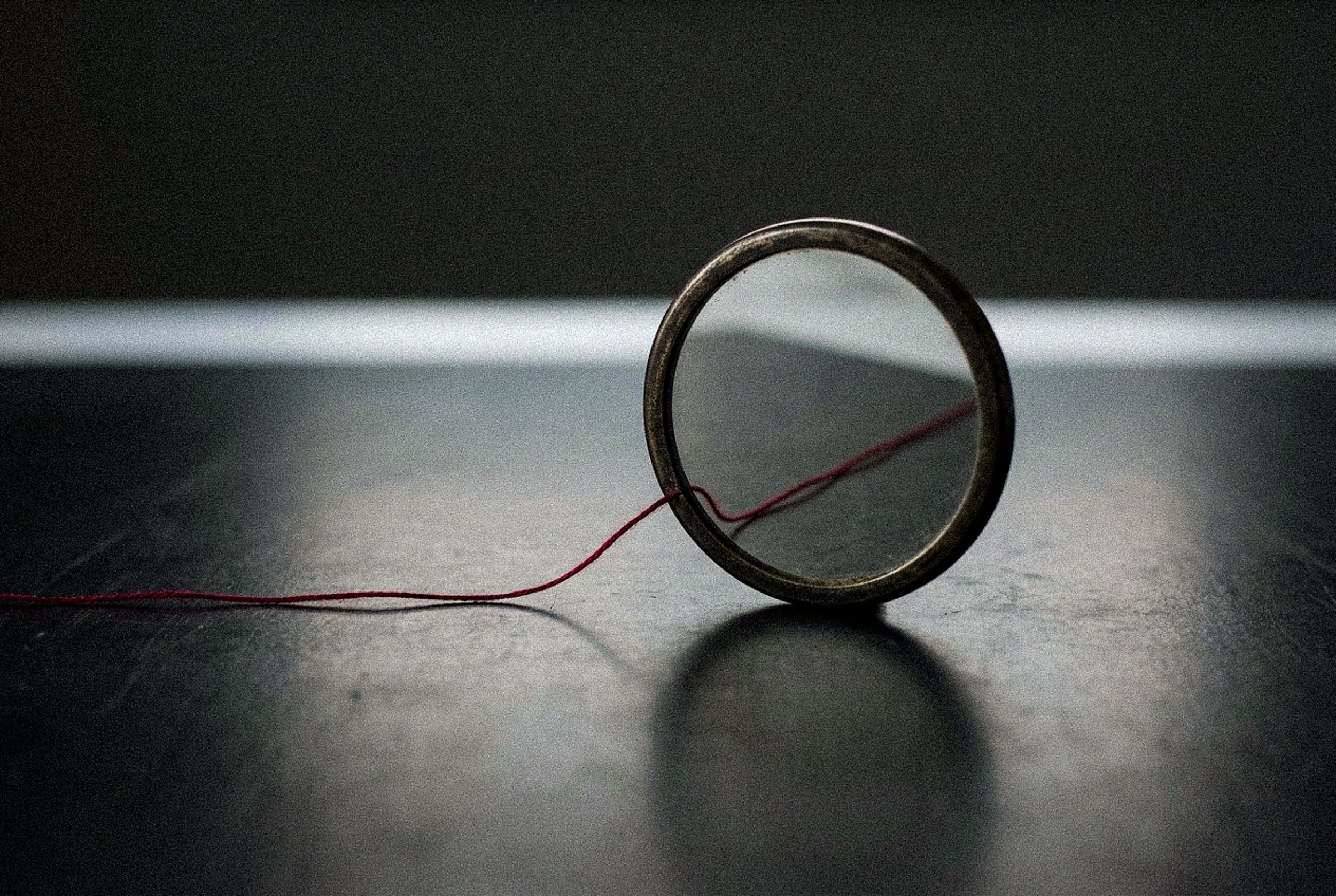 An antique round mirror stands upright on a dark surface, a fine red thread entering its frame and continuing inside the reflection, against a soft band of light.