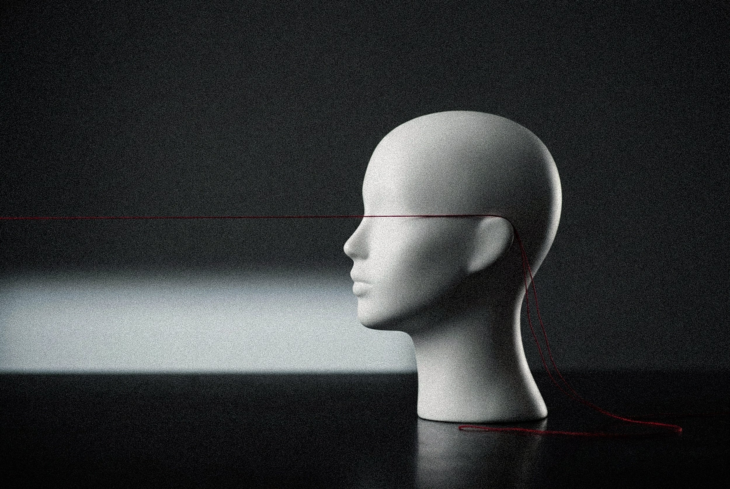 A smooth white mannequin head in profile on a dark surface, a fine red thread running straight across where the eyes would be, its loose ends falling behind the ear.