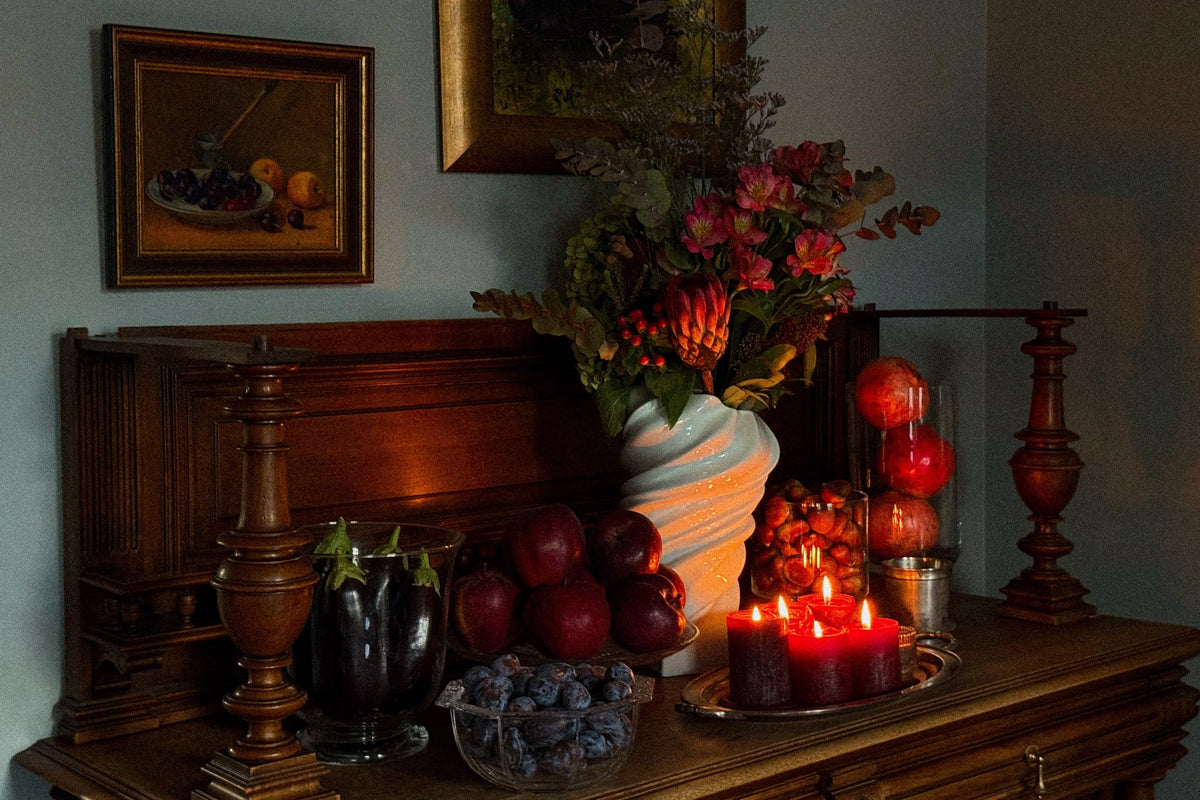 A fall still life on a dark wooden sideboard: a bouquet of alstroemerias and protea in a white twisted vase, red apples, plums in crystal, aubergines, pomegranates, and lit red candles on a silver tray.